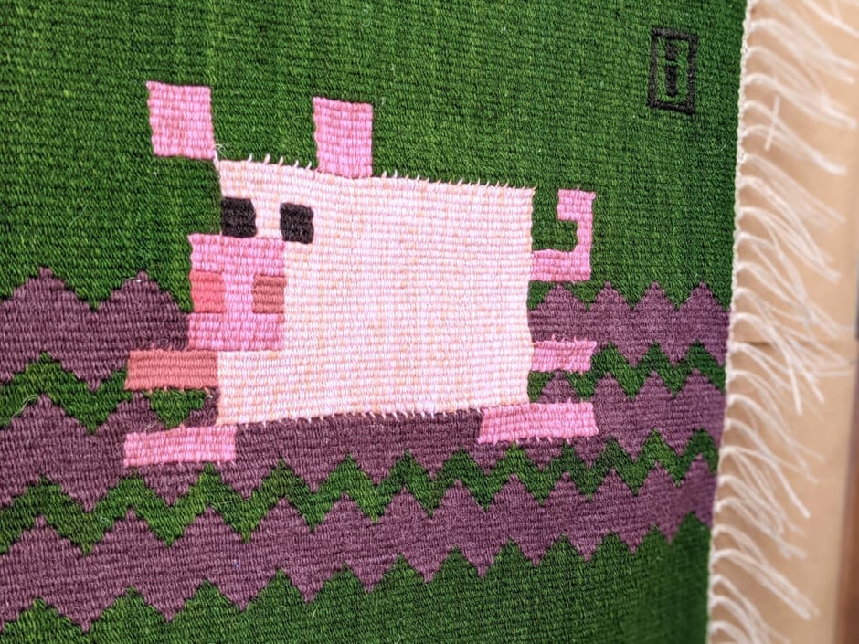 Un kilim-tenture murale en tissage au fond vert et un petit cochon rose jouant dans la boue. Dessin du tissage mural géométrisé.