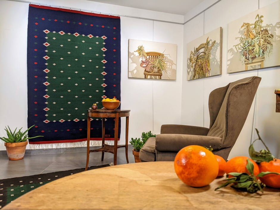 Intérieur avec, sur le mur, un grand kilim bleu-vert avec le motif de diamants, tissé à la main en laine. Devant, deux tables avec, posés sur eux, les oranges et les pommes, à droit un grand fauteuil voltaire et sur le mur trois tableaux - nature morte.
