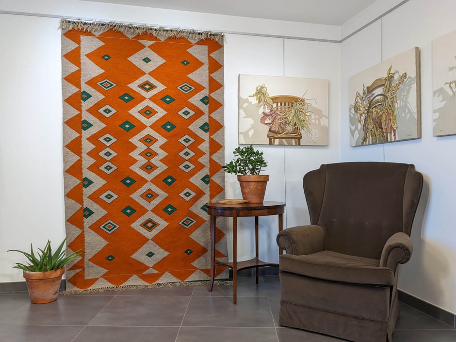 Intérieur avec, sur le mur, un grand kilim gris-orange, en zigzag et diamants. À droite, une table avec une plante succulente en pot de terre cuite, devant un grand fauteuil voltaire et sur le mur au-dessus, deux tableaux - nature morte.