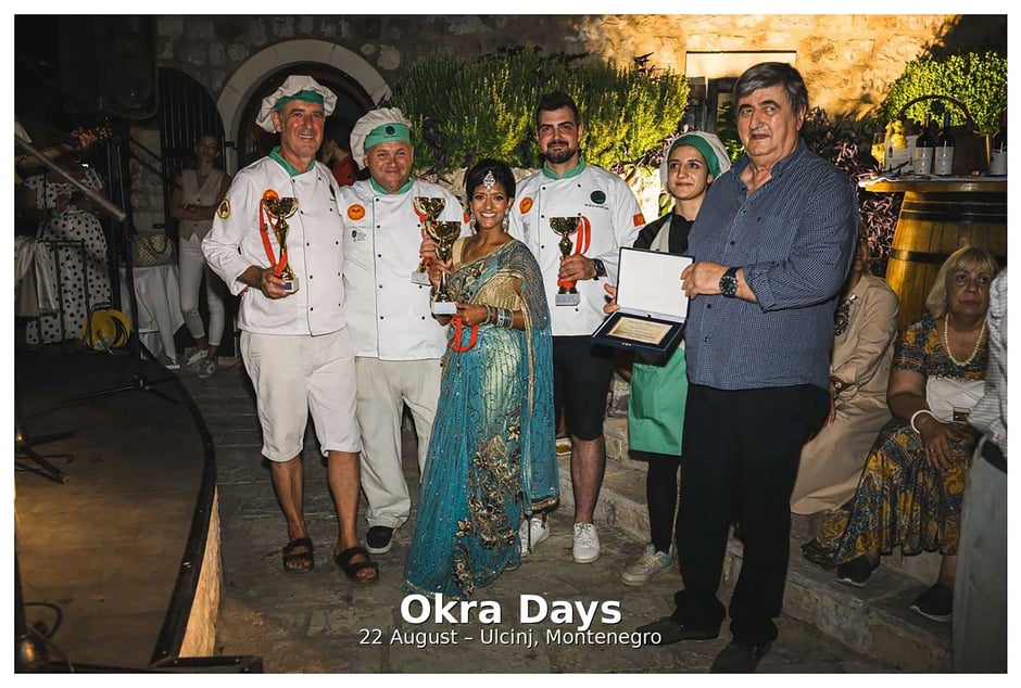 Group of chefs and participants holding trophies and awards during the Okra Days festival in Ulcinj, Montenegro, standing in a courtyard at night with an audience in the background
