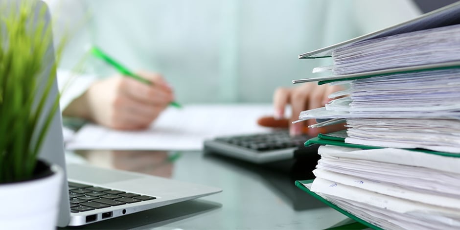 Große Stapel Akten auf einem Schreibtisch, im Hintergund ein Mitarbeiter oder eine Mitarbeiterin bei der Abrechnung.