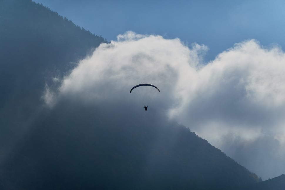 The image shows the photograph of a paraglider against the background of some sunlit white clouds and a dark mountain ridge..