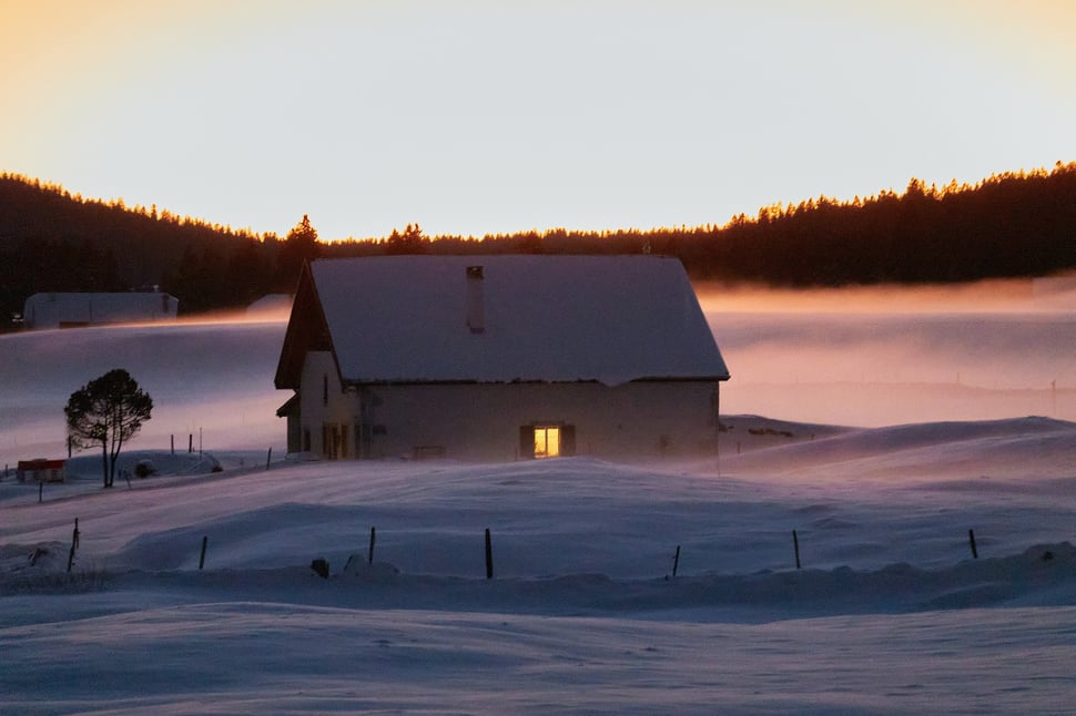 The image shows the photograph of a farm house in a Winter landscape of the Jura region (Switzerland). It is a very cold evening - the last sunlight illuminates the haze surrounding the house.