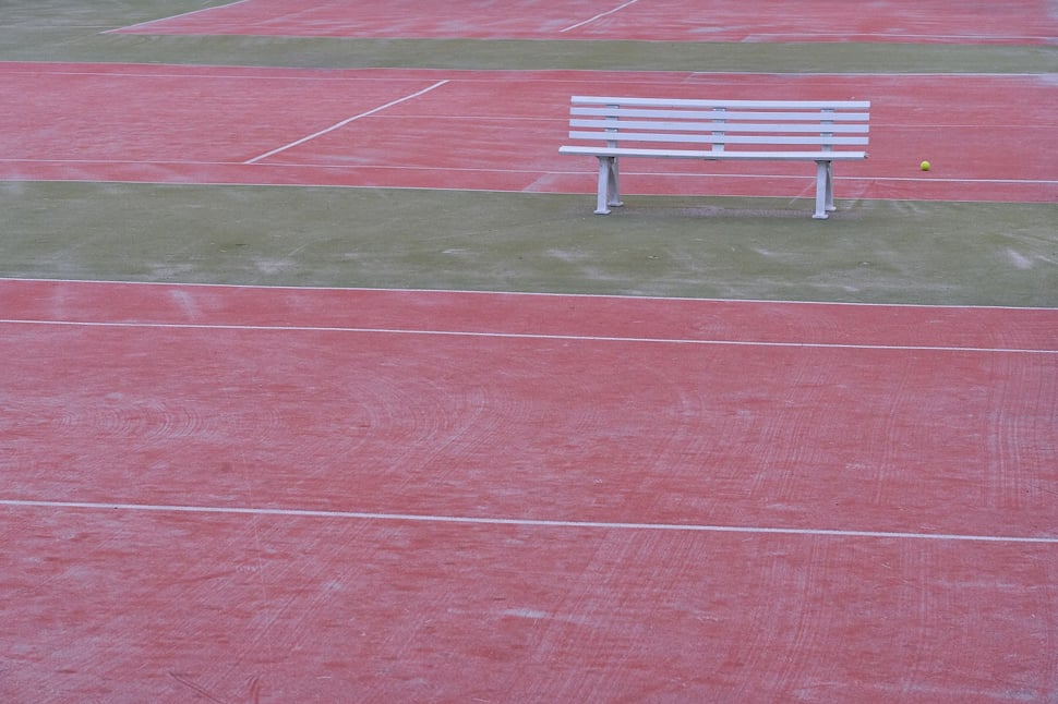The image shows the photograph of vacant tennis courts an a white bench and a single tennis ball. It is early morning.