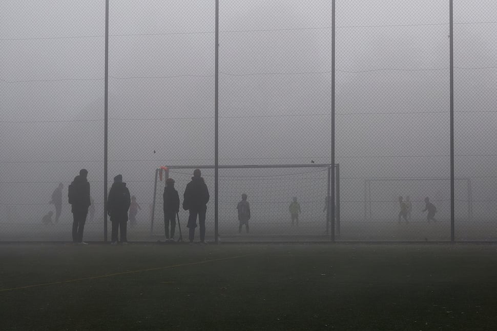 The image shows the photograph of kids playing soccer behind a high chainlink fence while their parents watch. It is a foggy Winter morning.