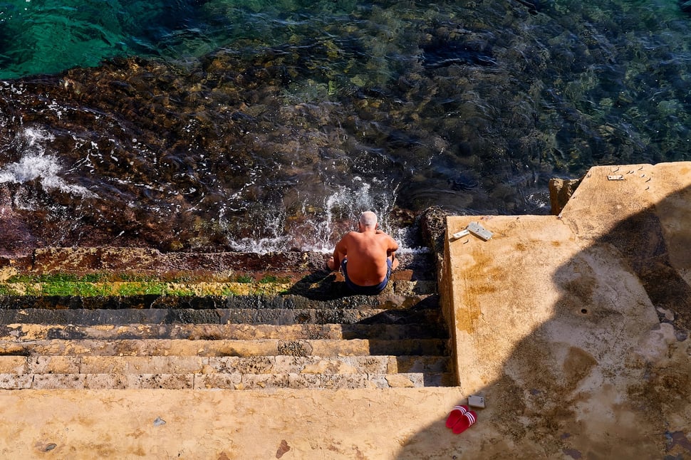 The image shows the photograph of an old man in swimming trunks sitting on the rocky steps of a sea bath in Santa Cesare Terme (Apulia). It is morning and everything is bathed in warm sunlight.