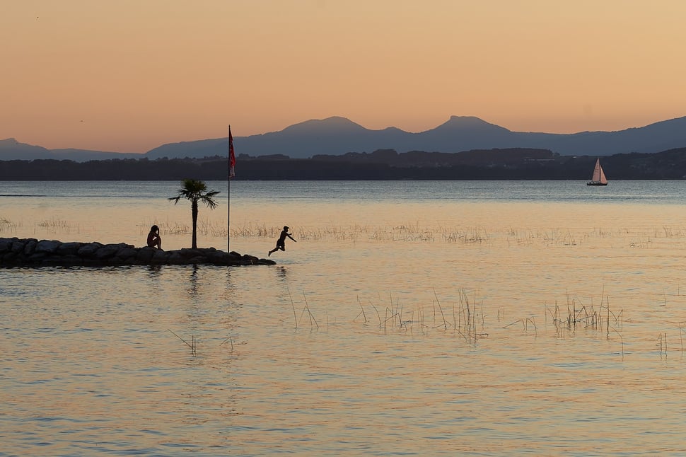 The image shows the photograph of a boy jumping into the water of Lake Morat at dusk while a second person is sitting on the shore and watching. There are a sailboat and mountains in the background.