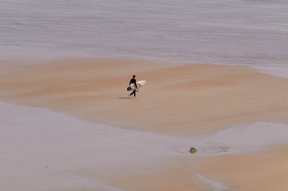 The image shows the minimalistic photography of a surfer carrying his surf board on a beach in Finistère (Brittany).