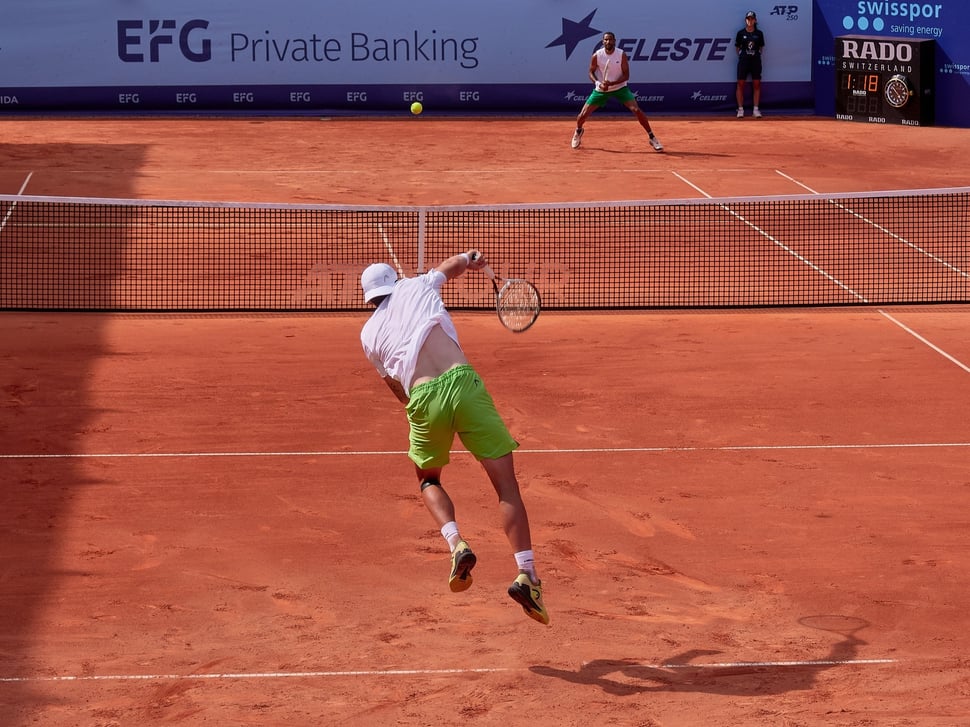 The image shows the photograph of the tennis player Jérôme Kym serving (from behind) at the Swiss Open 2025 in Gstaad. You can see his opponent - Calvin Hemery - getting ready to return.