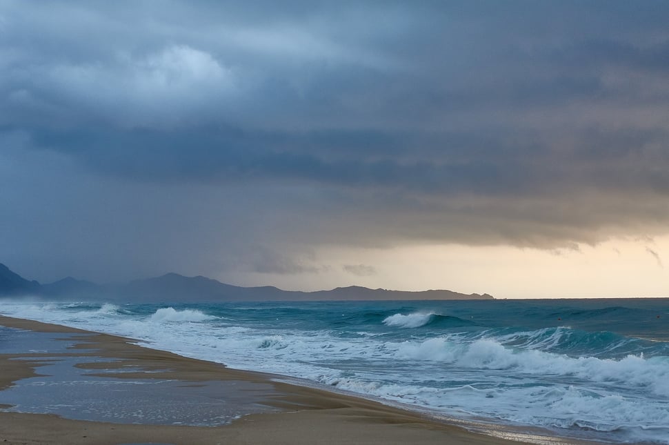 The image shows the photograph of a beach in Sardinia on a fall morning. Dark clouds and a range of mountains are visible in the background. 