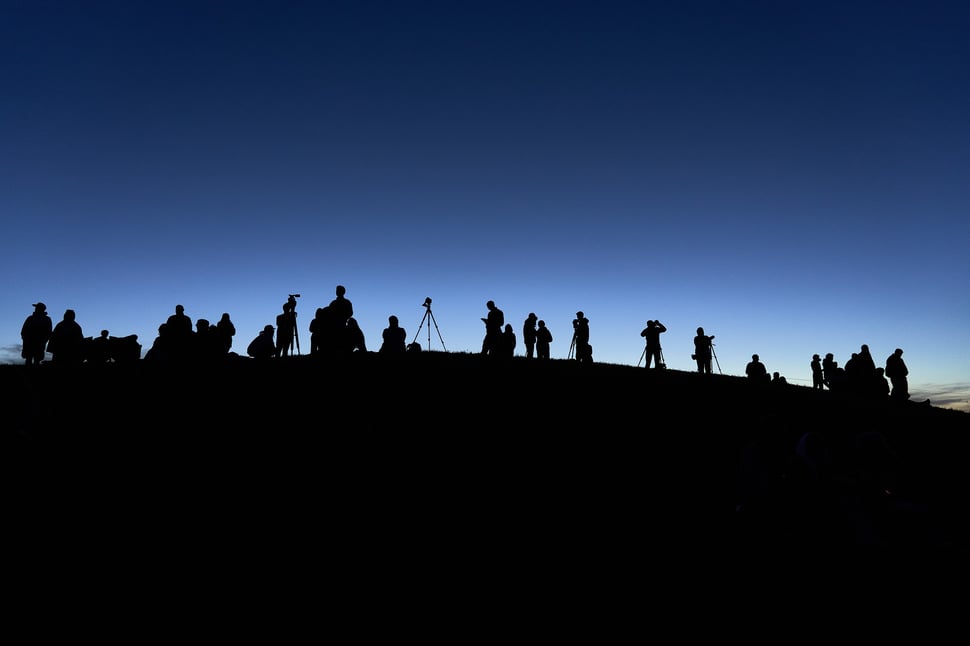 The image shows the photograph - shot against the last light of the day - of black shapes of people on a hill with photograhic equipmen against the background of a blue sky. 