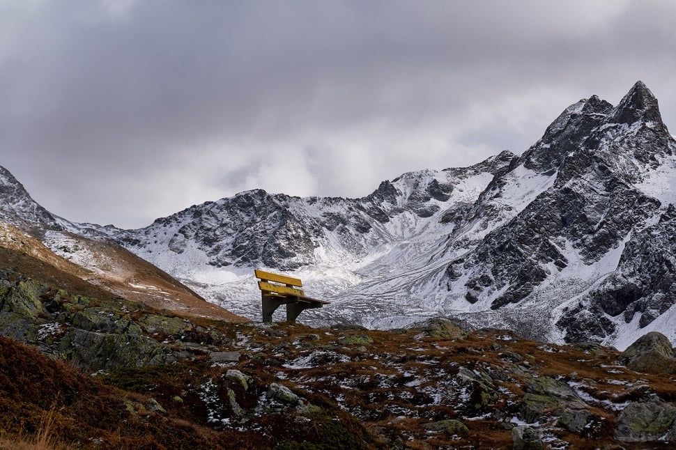 The image shows the photograph of a yellow bench in a mountainous setting under a grey sky. The location is Muottas Muragl (Engadin).