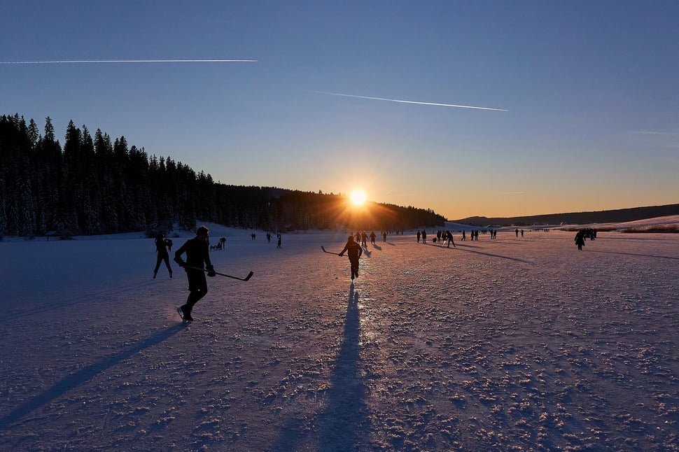The image shows the photograph of a lot of people walking or ice skating or playing ice hockey on a frozen lake while the sun is setting. The lake is called Lac des Taillères, the location is La Brévine (Jura region).